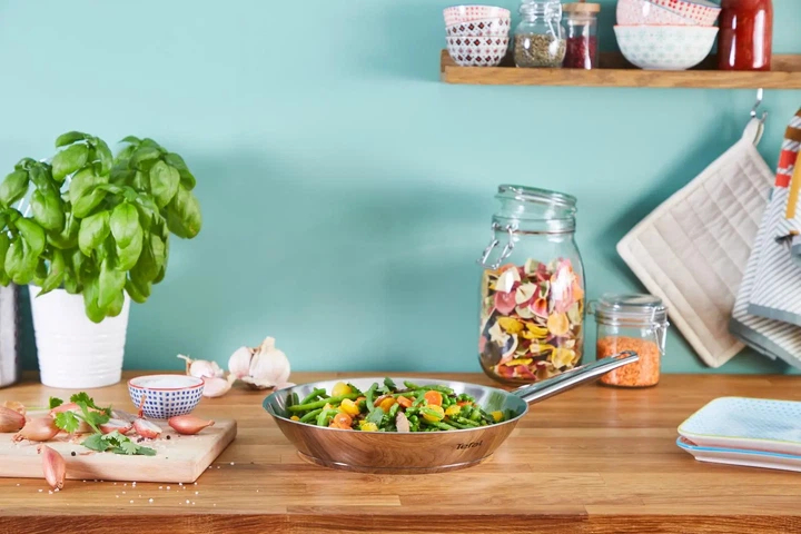 Drewniany stół z produktami do gotowania: bazylia, szalotka, czosnek, przyprawy i patelnia z warzywami.