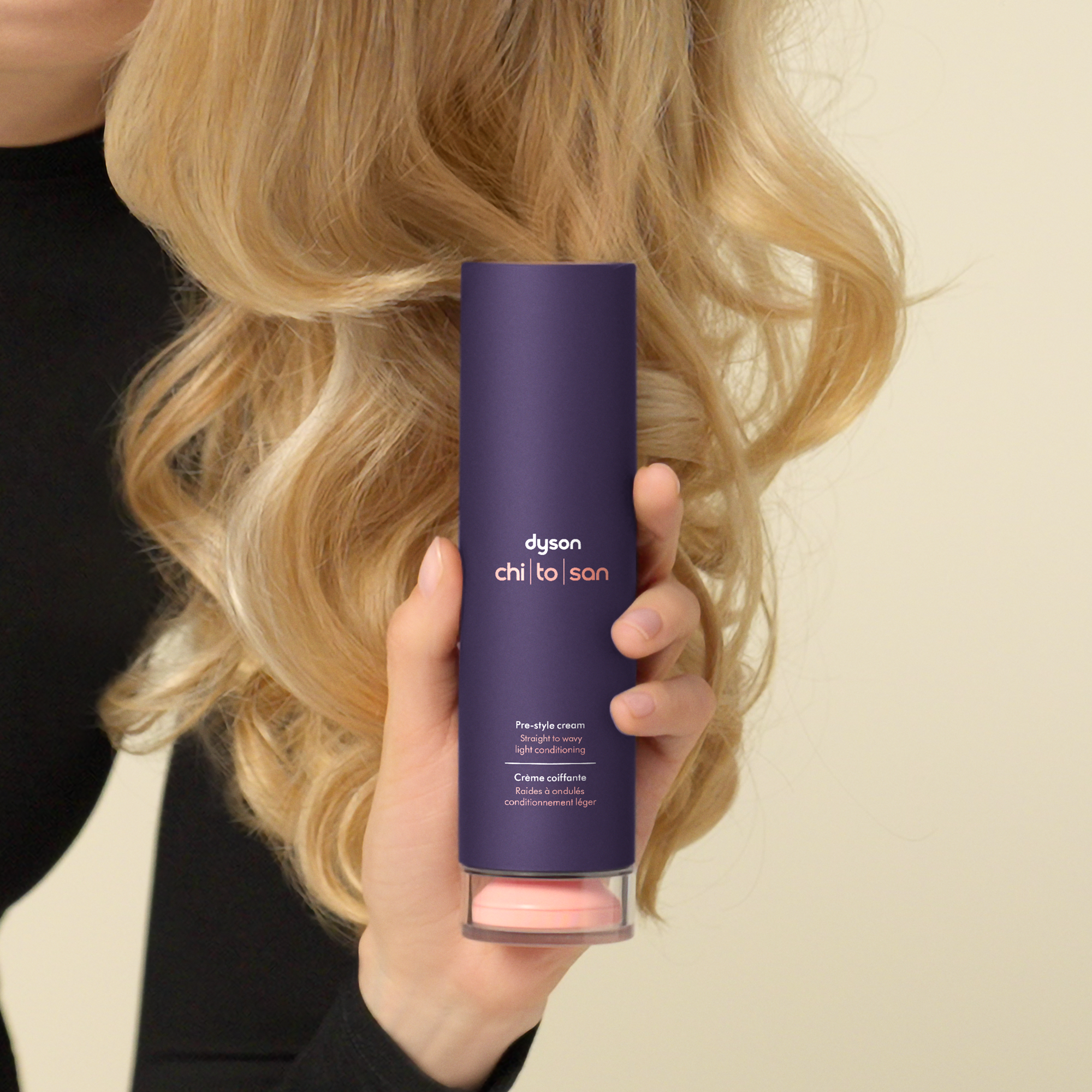 Eine Hand hält eine lila Tube Dyson Haarpflegecreme vor blondes, welliges Haar.