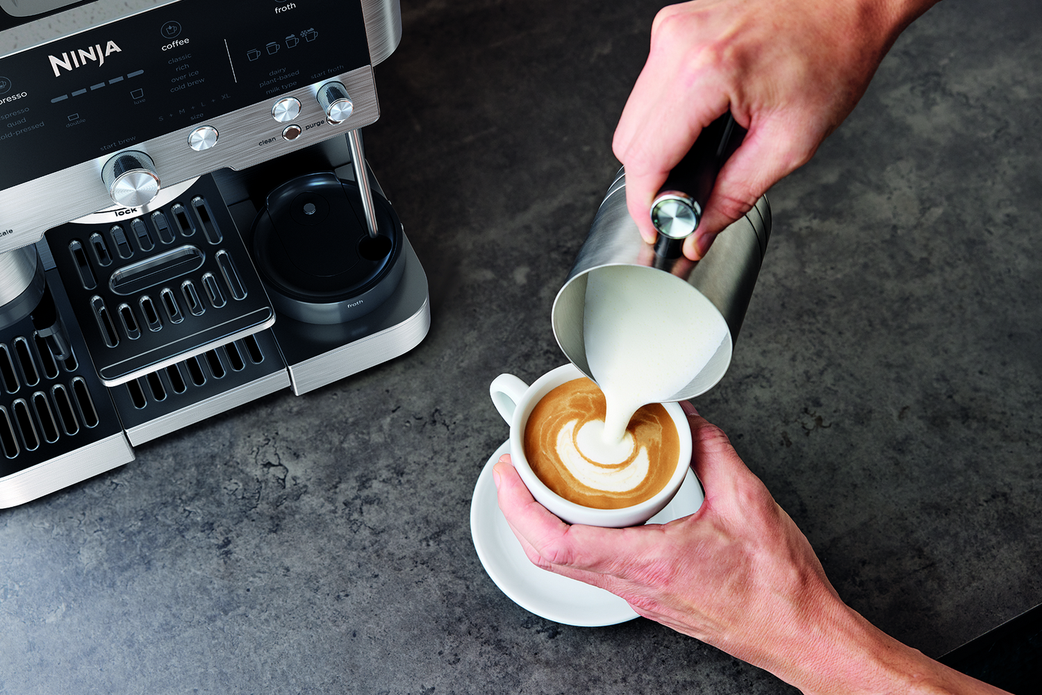 Una mano vierte espuma de leche en una taza de café junto a una cafetera.
