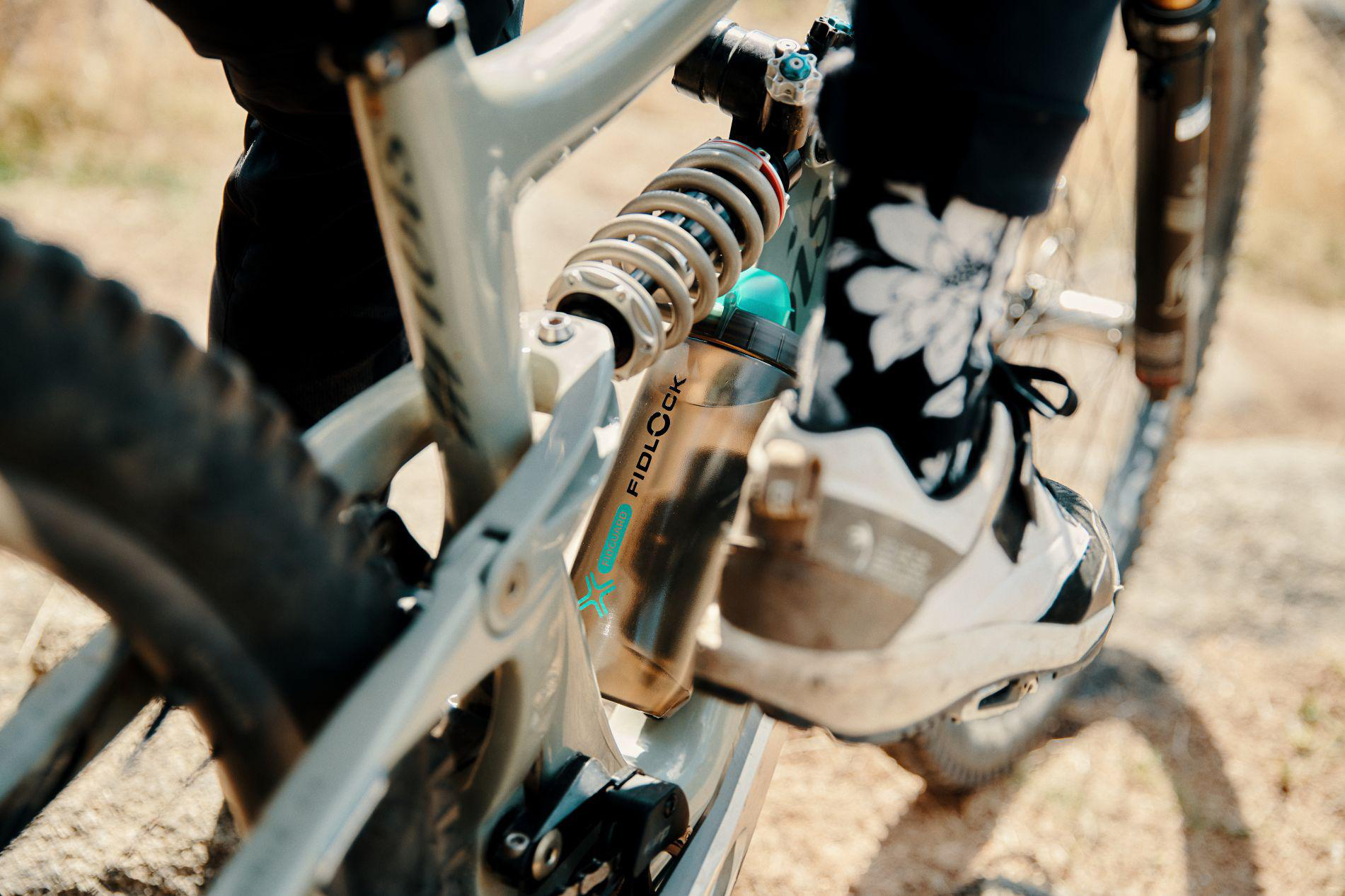 Nahaufnahme einer Fahrradflasche am Mountainbike-Stoßdämpfer mit einem Radfahrerfuß in gemusterten Socken.