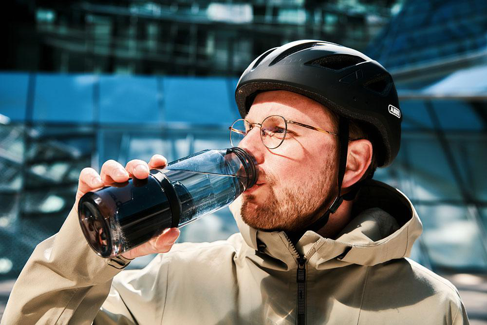 Mann mit Helm trinkt aus Flasche vor modernem Gebäude.