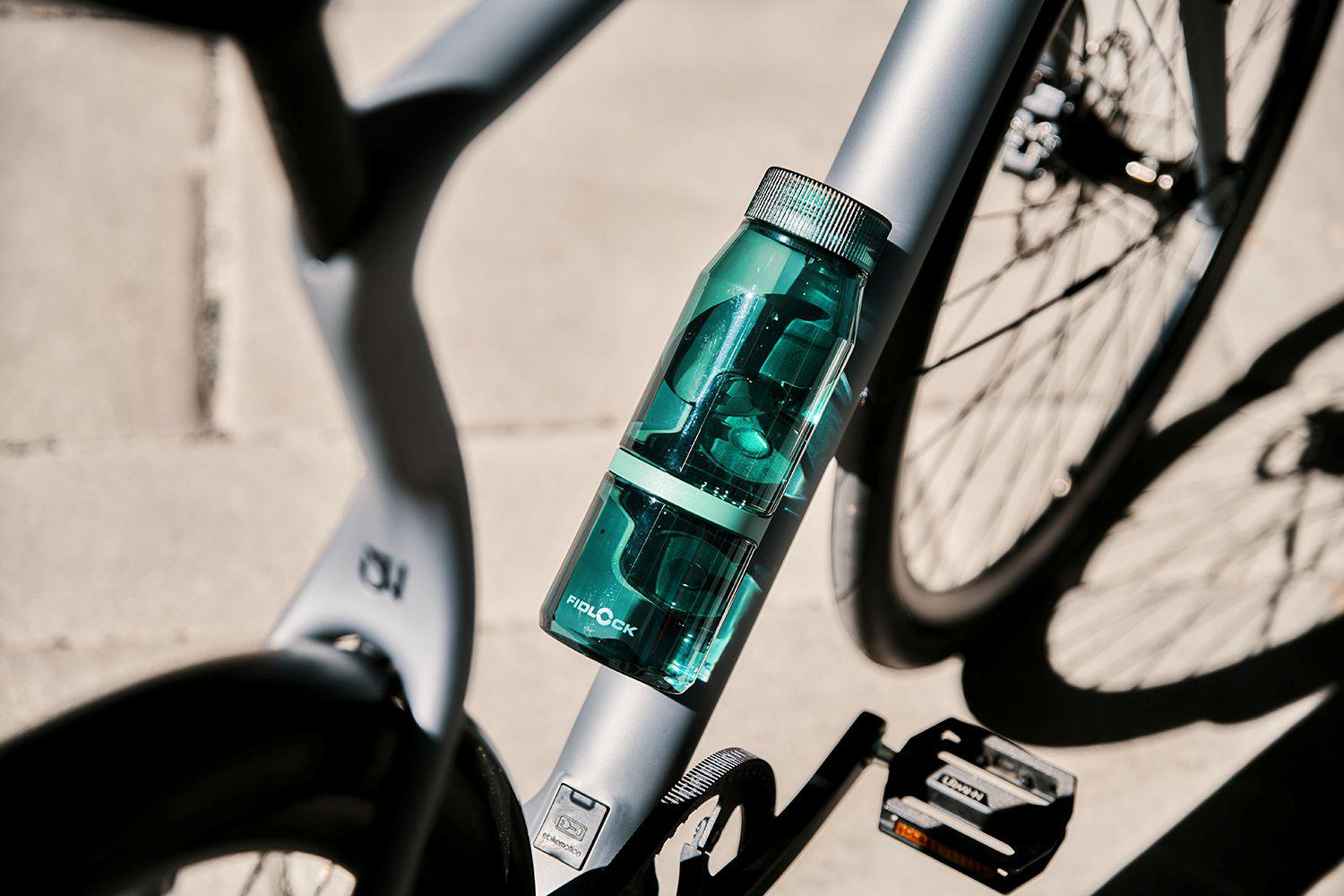 Eine grüne Wasserflasche ist am grauen Fahrradrahmen befestigt, im Hintergrund ein Rad.