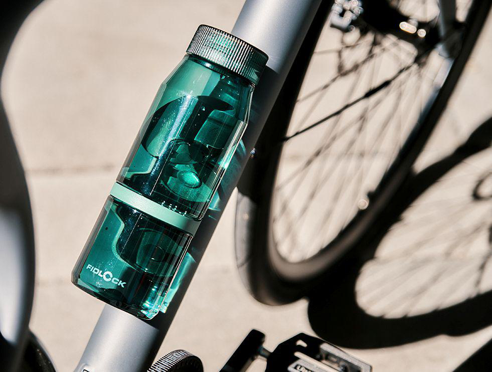 Grüne FIDLOCK-Wasserflasche an einem Fahrradrahmen, Rad im Hintergrund.