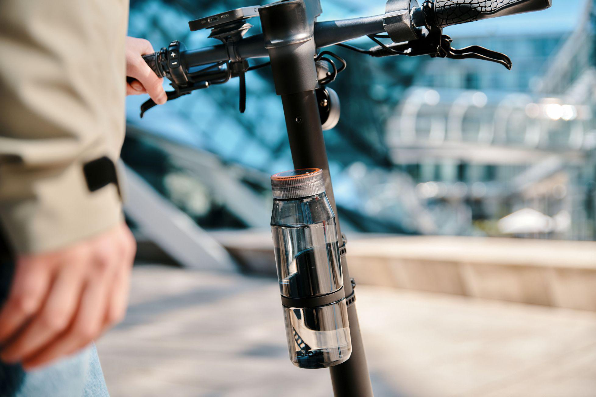 Person hält Lenker eines E-Scooters mit befestigter Wasserflasche. Hintergrund unscharf.