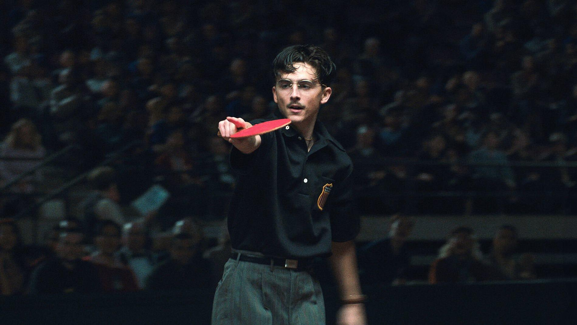 Ein Mann mit Brille und Schnurrbart hält einen roten Tischtennisschläger und zeigt darauf.