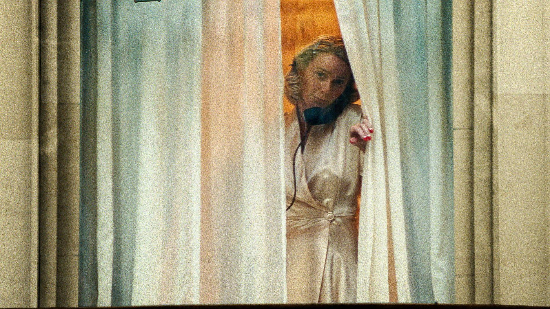 Eine Frau in einem seidenen Kleid blickt hinter Vorhängen hervor.