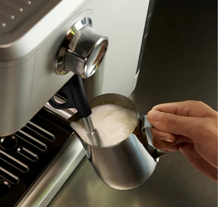 Dłoń trzyma metalowy dzbanek z mlekiem spienianym przez ekspres do kawy.
