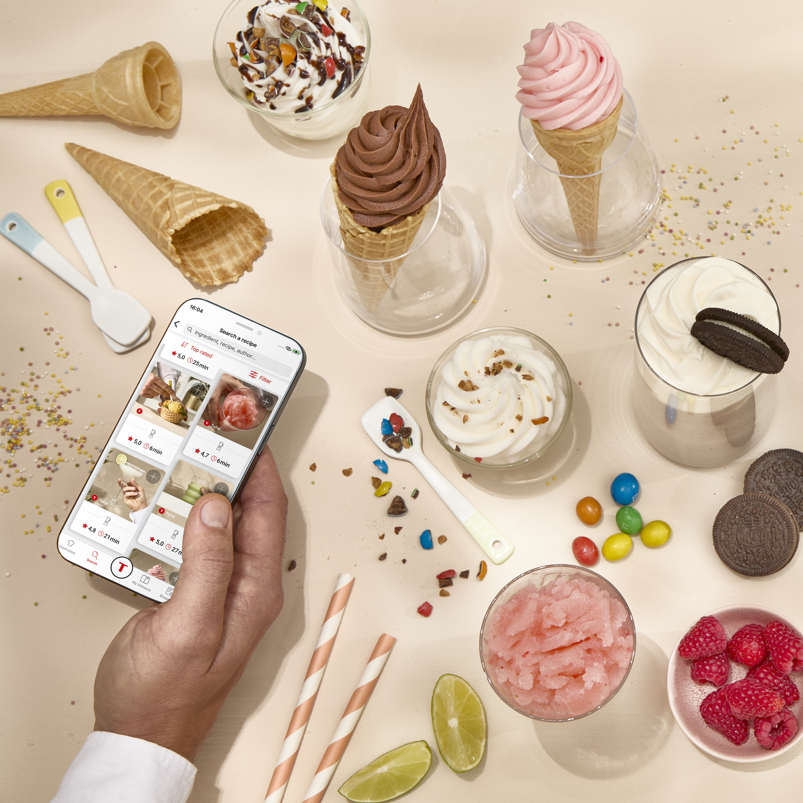 Eine Hand hält ein Smartphone mit Rezepten, umgeben von Eis, Waffeln und Süßigkeiten.