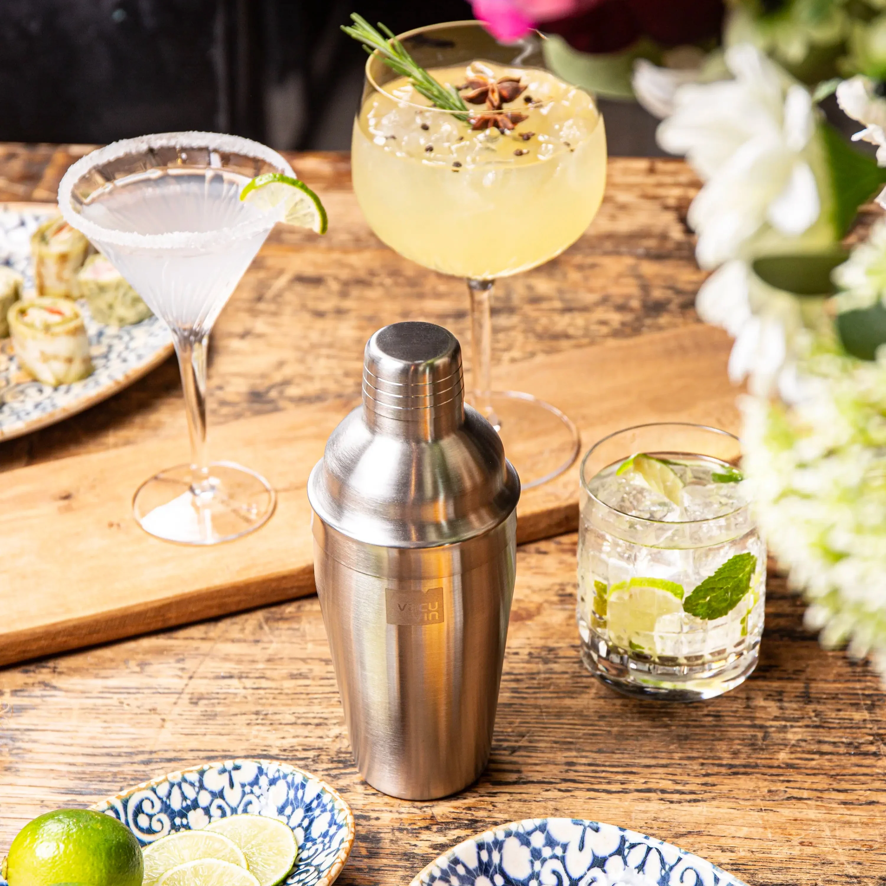 Cocktailshaker mit zwei Cocktails, garniert mit Limette und Kräutern, und Sushi-Röllchen.