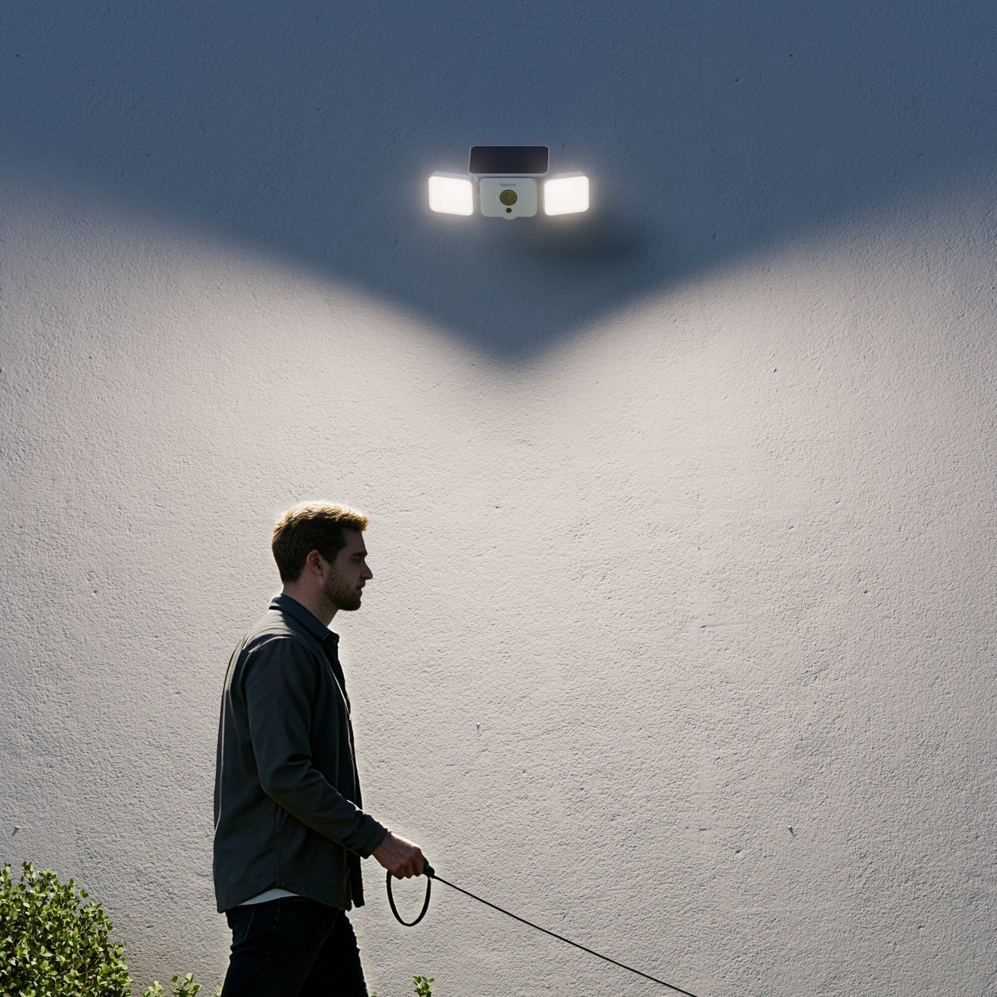Ein Mann geht mit einem Hund an einer Wand entlang, beleuchtet von einem Scheinwerfer.