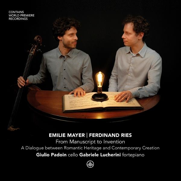 Zwei Männer sitzen an einem Tisch, einer spielt Cello. Noten und Lampe sind sichtbar.