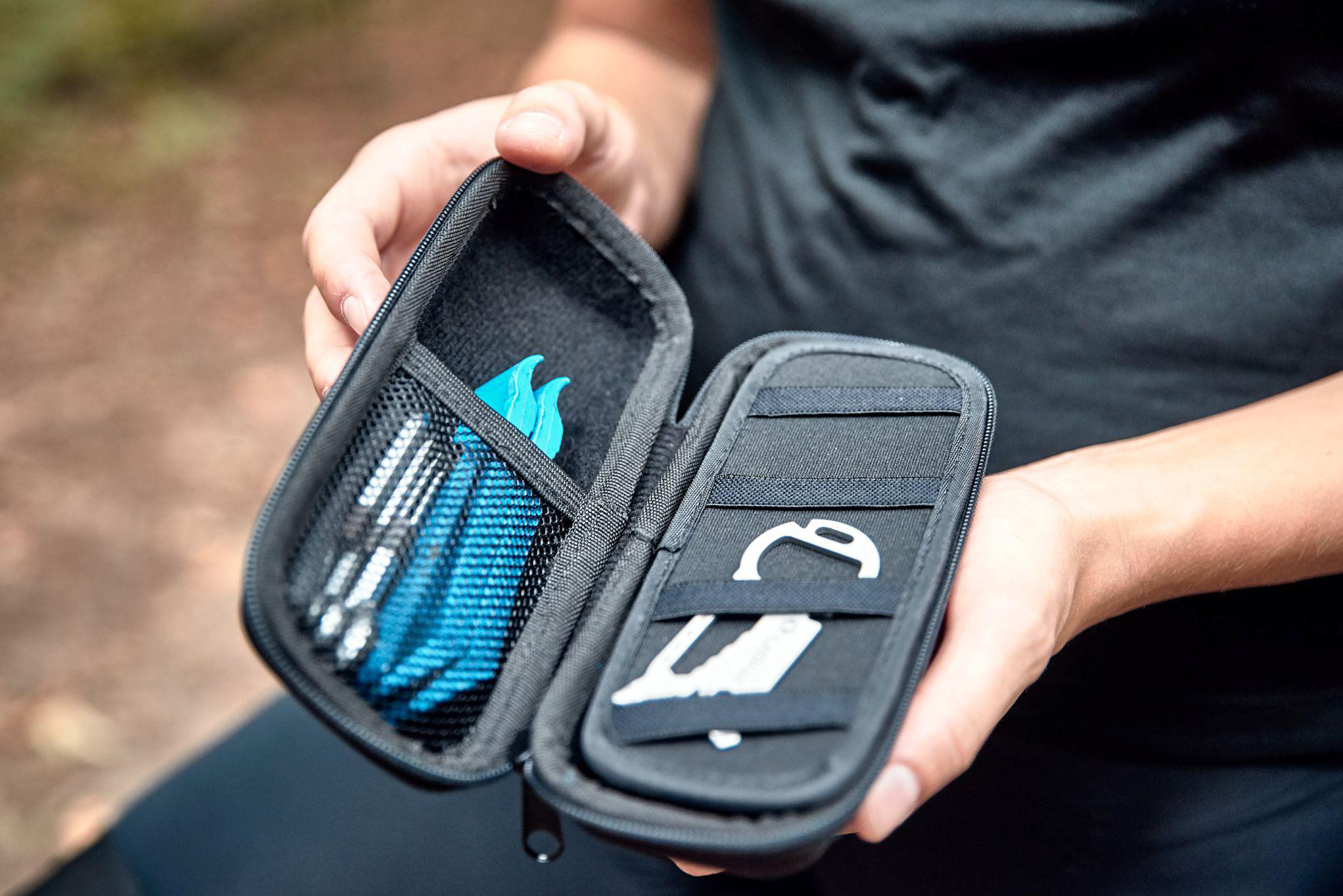 Eine Person öffnet eine kleine schwarze Tasche mit Werkzeugen, darunter blaue Reifenheber und silberne Werkzeuge.