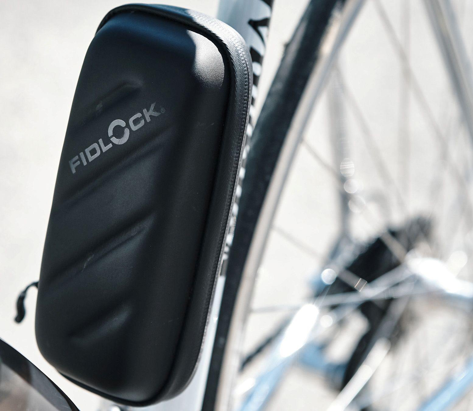Schwarze Fahrradtasche mit 'FIDLOCK'-Logo, befestigt am Fahrradrahmen, Rad im Hintergrund.