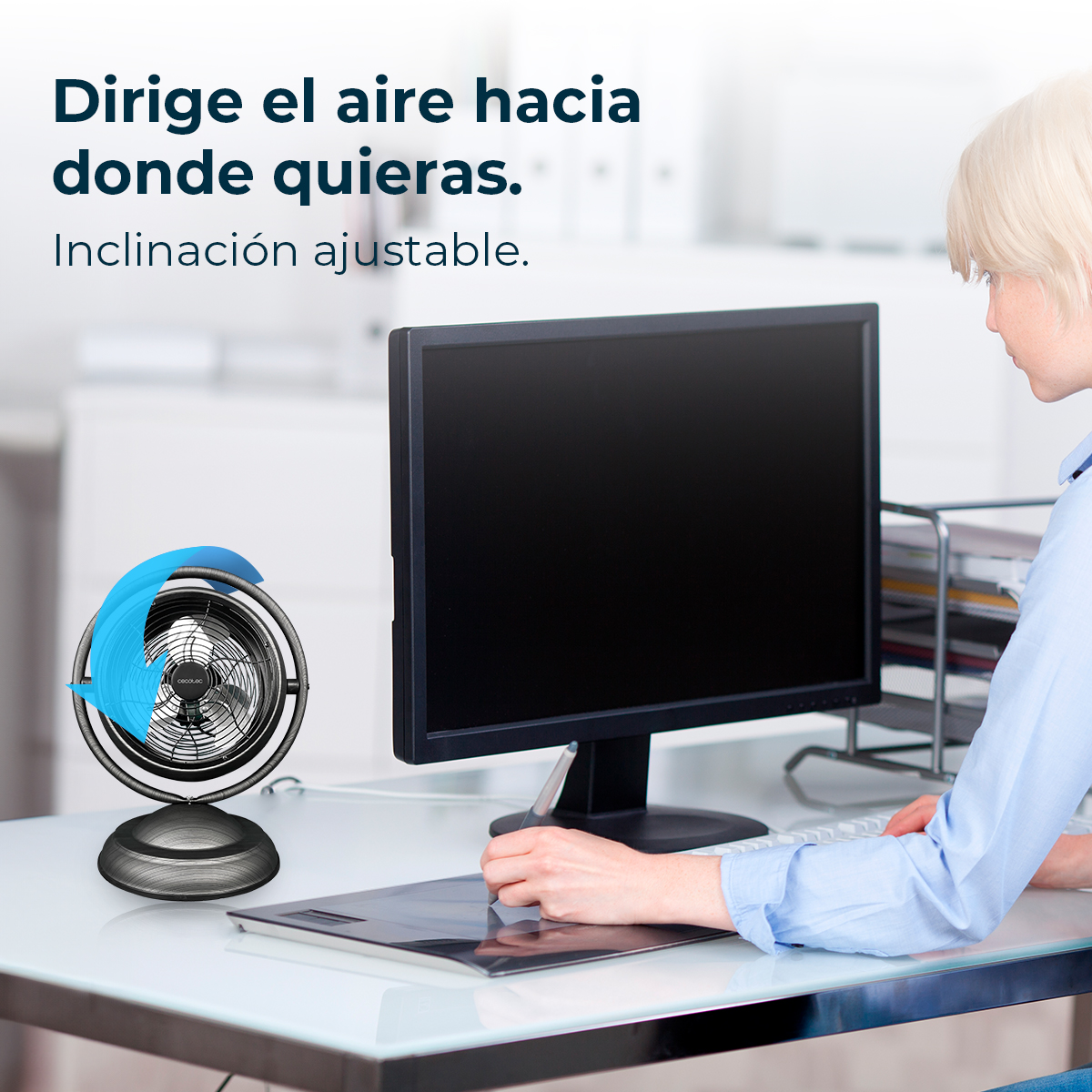Un ventilador pequeño y un monitor de computadora en un escritorio, una mujer trabaja.
