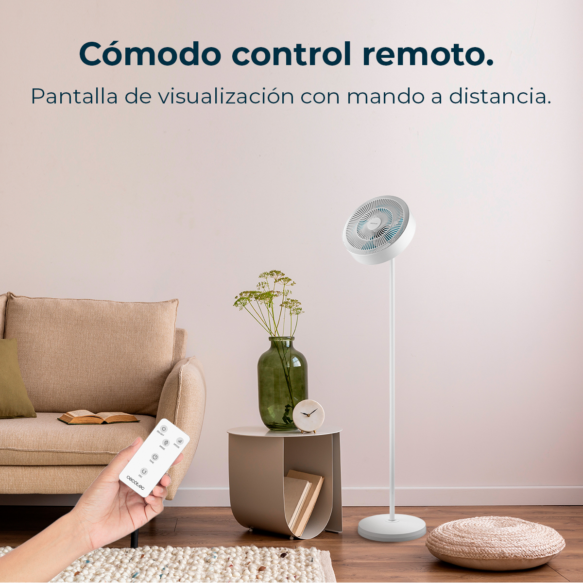 Un ventilador de pie blanco junto a una mesita auxiliar con jarrón y libro. Una mano sostiene un mando.