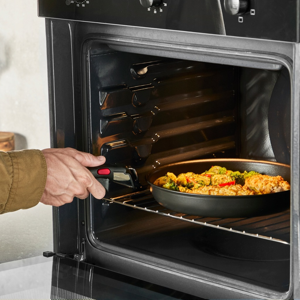 Ręka wkłada patelnię z kurczakiem i warzywami do otwartego piekarnika.