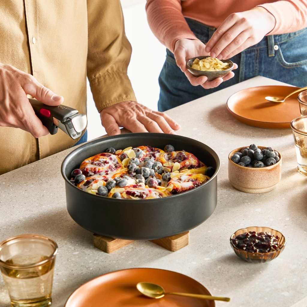 Patelnia z ciastem jagodowym na blacie, ręce dodają polewy.