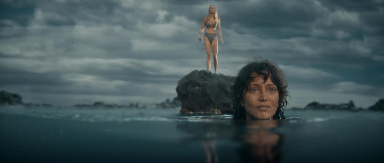 Eine Frau im Wasser blickt nach oben, eine andere steht auf einem Felsen.