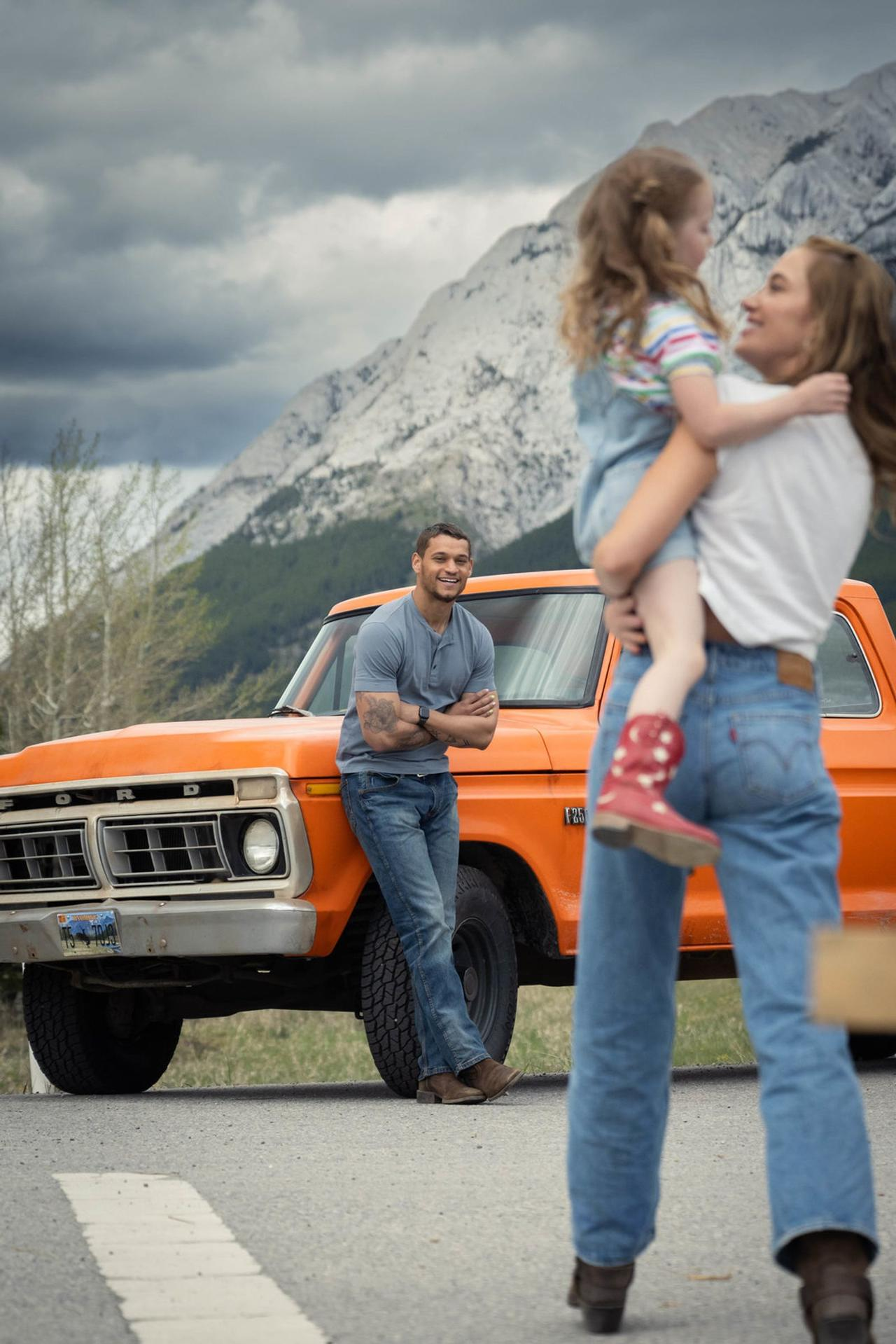 Mann lehnt an orangefarbenem Ford-Truck, Frau hält Kind. Berge im Hintergrund.