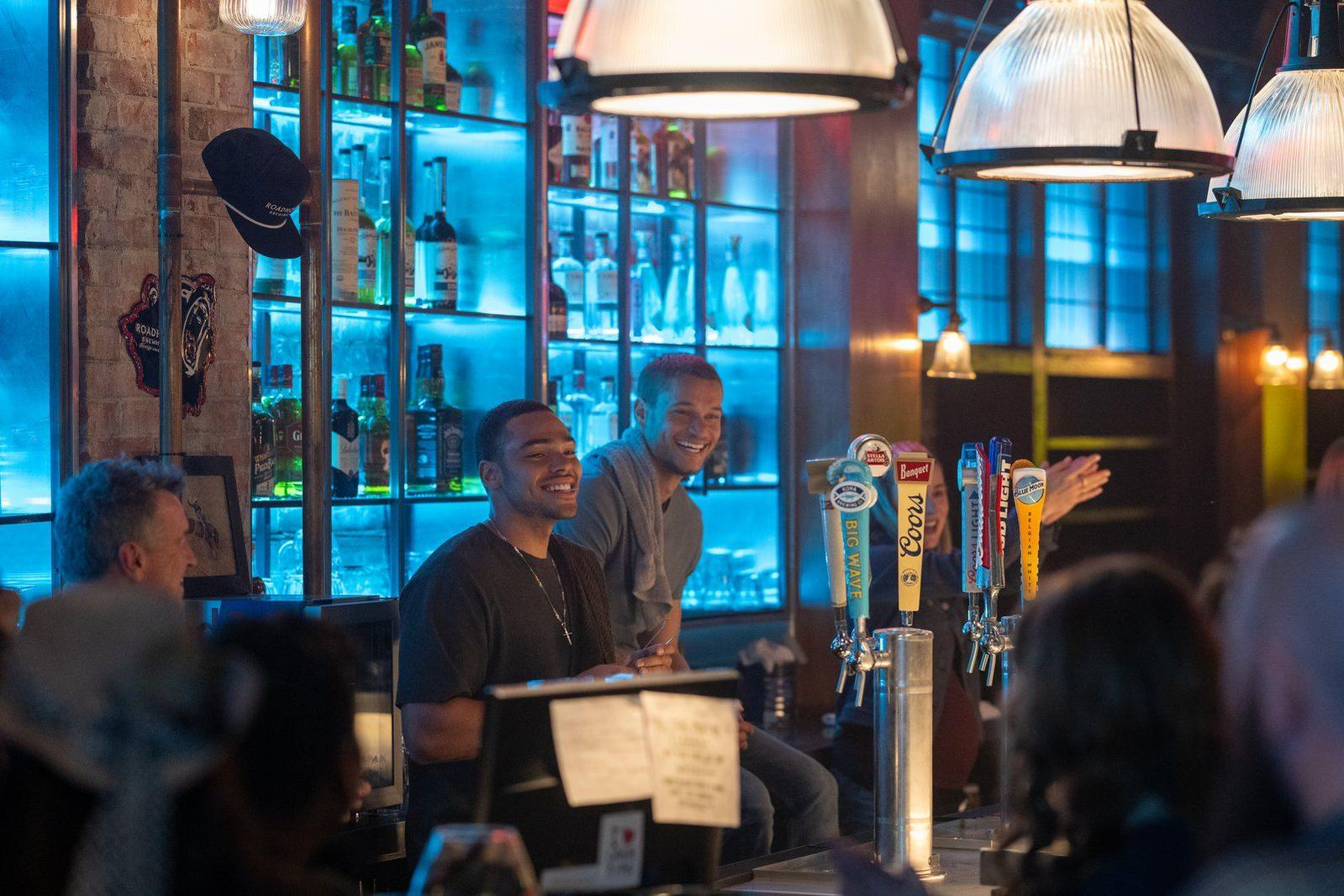 Zwei Männer lachen hinter einer Bar mit Bierhähnen und beleuchteten Regalen.