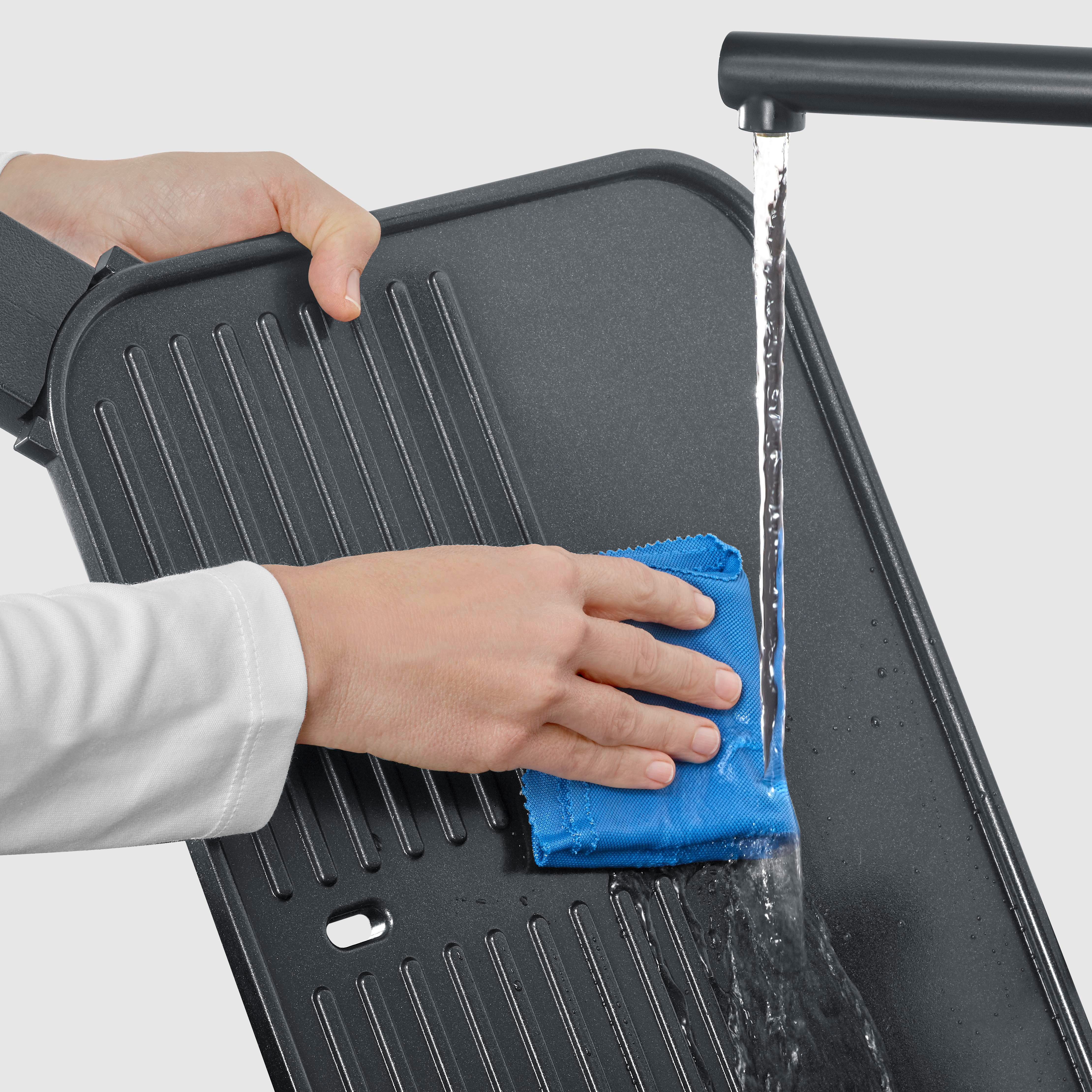 Hände reinigen eine Grillplatte unter fließendem Wasser mit einem blauen Tuch.