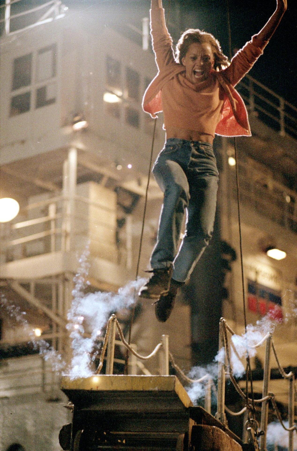 Eine Person springt mit erhobenen Armen, Rauch steigt von unten auf. Industrielles Gebäude im Hintergrund.