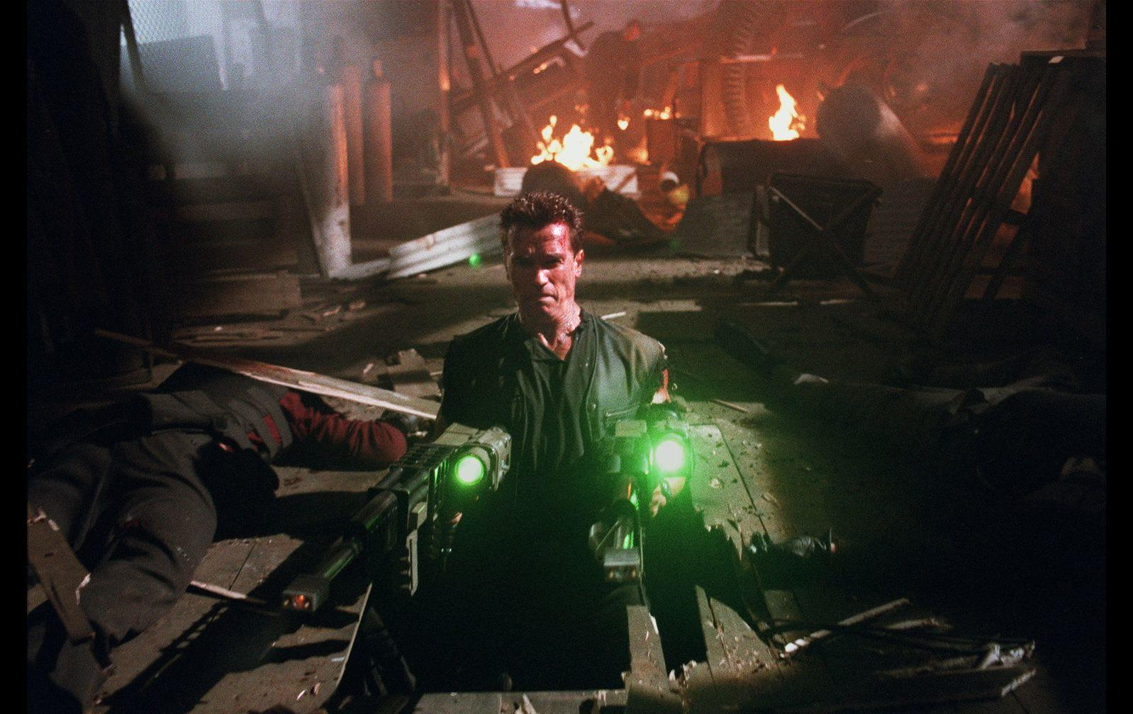 Arnold Schwarzenegger mit grünen Laserwaffen in einem brennenden Raum mit Leichen.