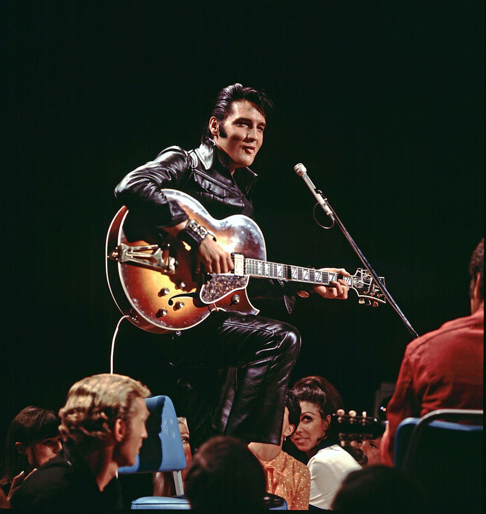 Elvis Presley in schwarzer Lederkleidung spielt Gitarre vor Publikum.