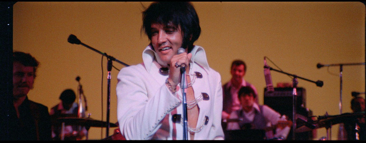 Elvis Presley in weißem Anzug singt ins Mikrofon, Band im Hintergrund.