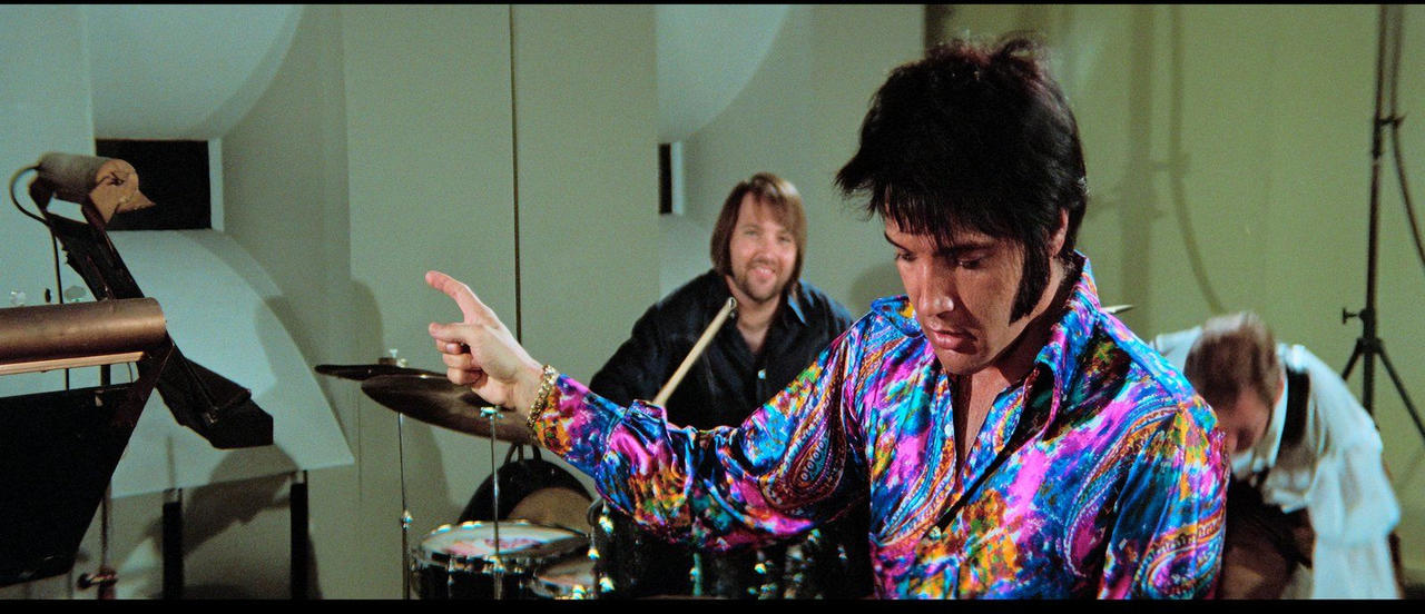 Elvis Presley in buntem Hemd dirigiert Musiker am Schlagzeug und Bass.