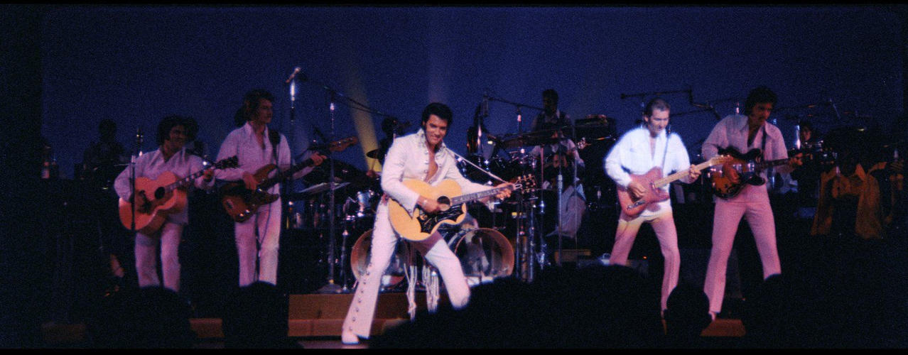 Elvis Presley mit Band auf der Bühne, spielt Gitarre in einem weißen Anzug.