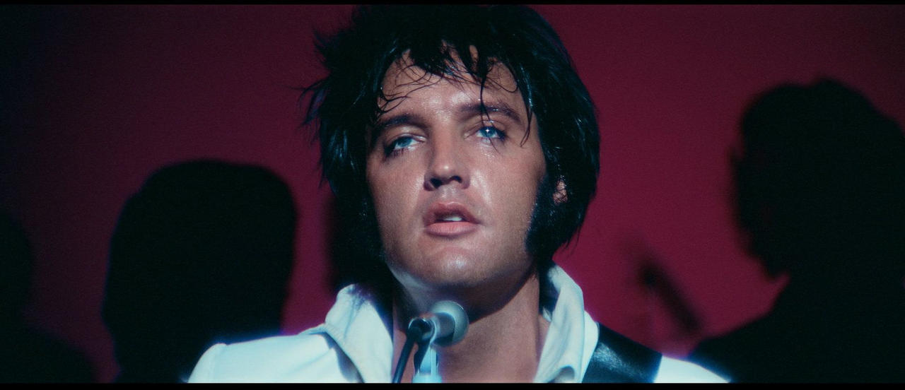Elvis Presley singt in einem weißen Hemd vor einem roten Hintergrund.
