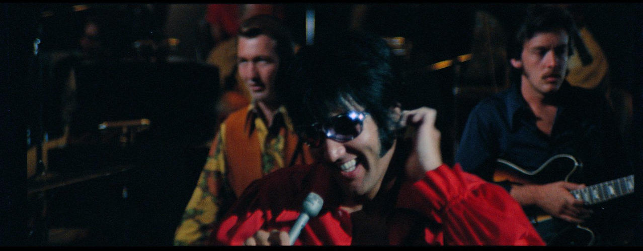 Elvis Presley in rotem Hemd mit Sonnenbrille singt, Band im Hintergrund.