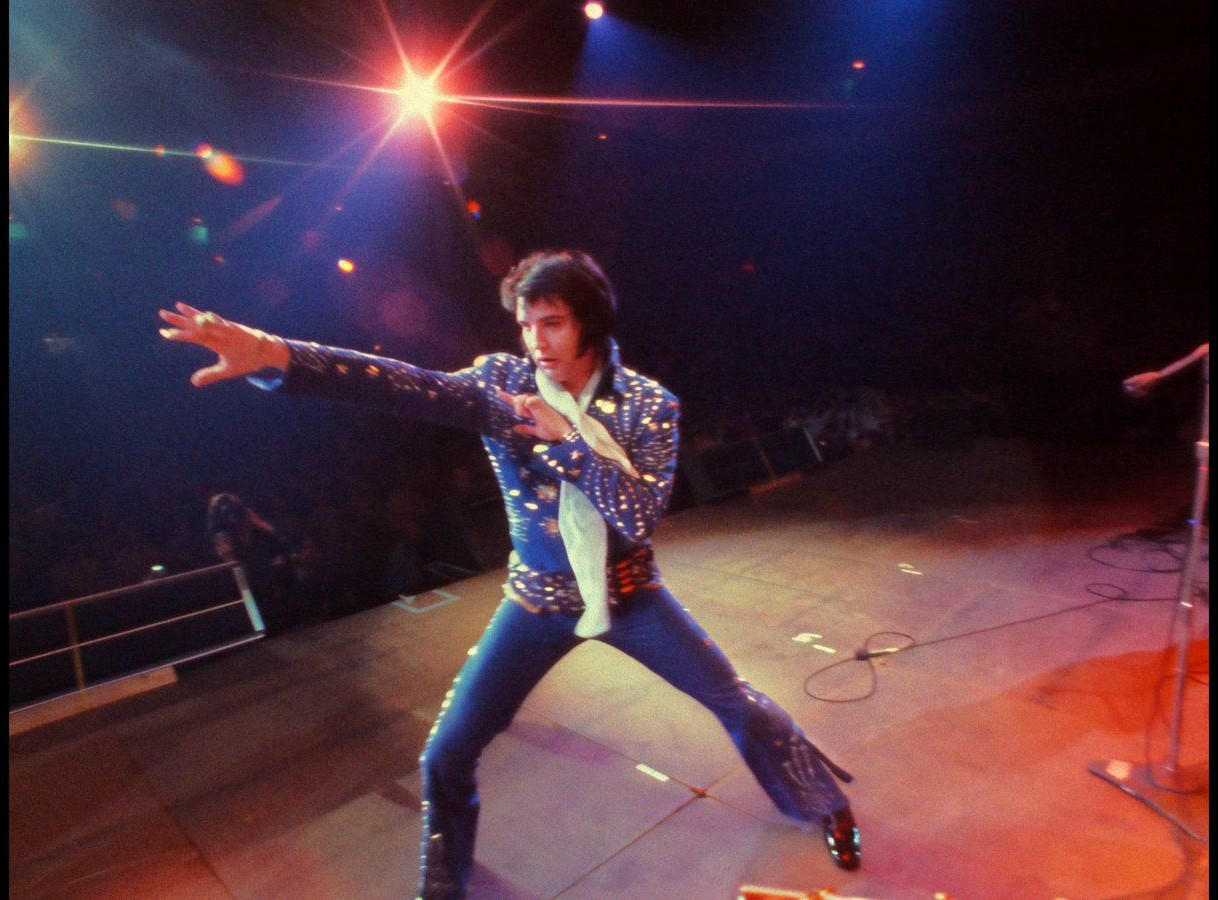 Elvis Presley in einem blauen Pailletten-Jumpsuit tritt mit ausgestreckten Armen auf.