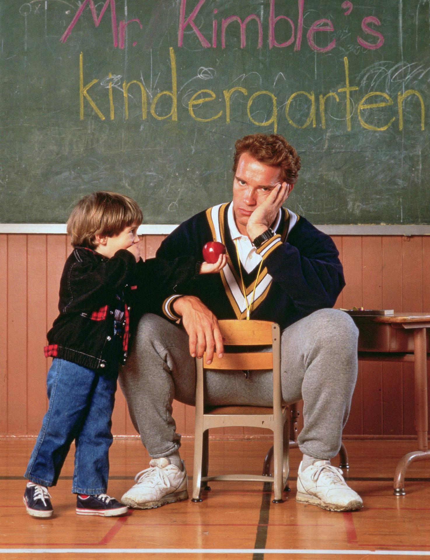 Ein Mann sitzt auf einem Stuhl vor einer Tafel mit "Mr. Kimble's Kindergarten".