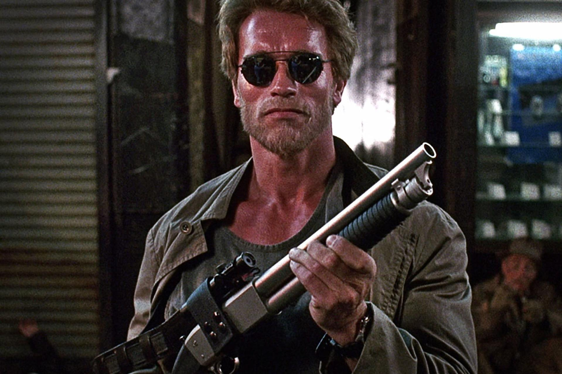 Ein Mann mit Sonnenbrille und Bart hält eine Schrotflinte, trägt eine Jacke.