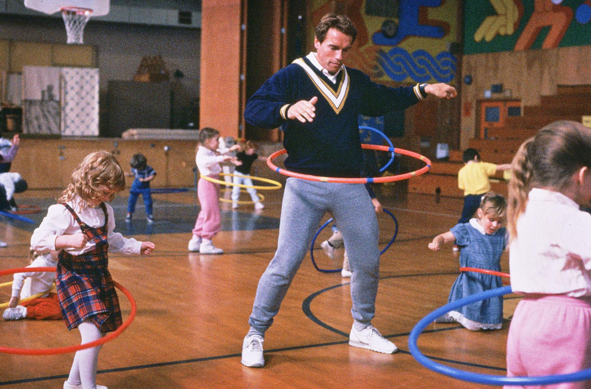 Arnold Schwarzenegger hulbert mit Kindern in einer Turnhalle.