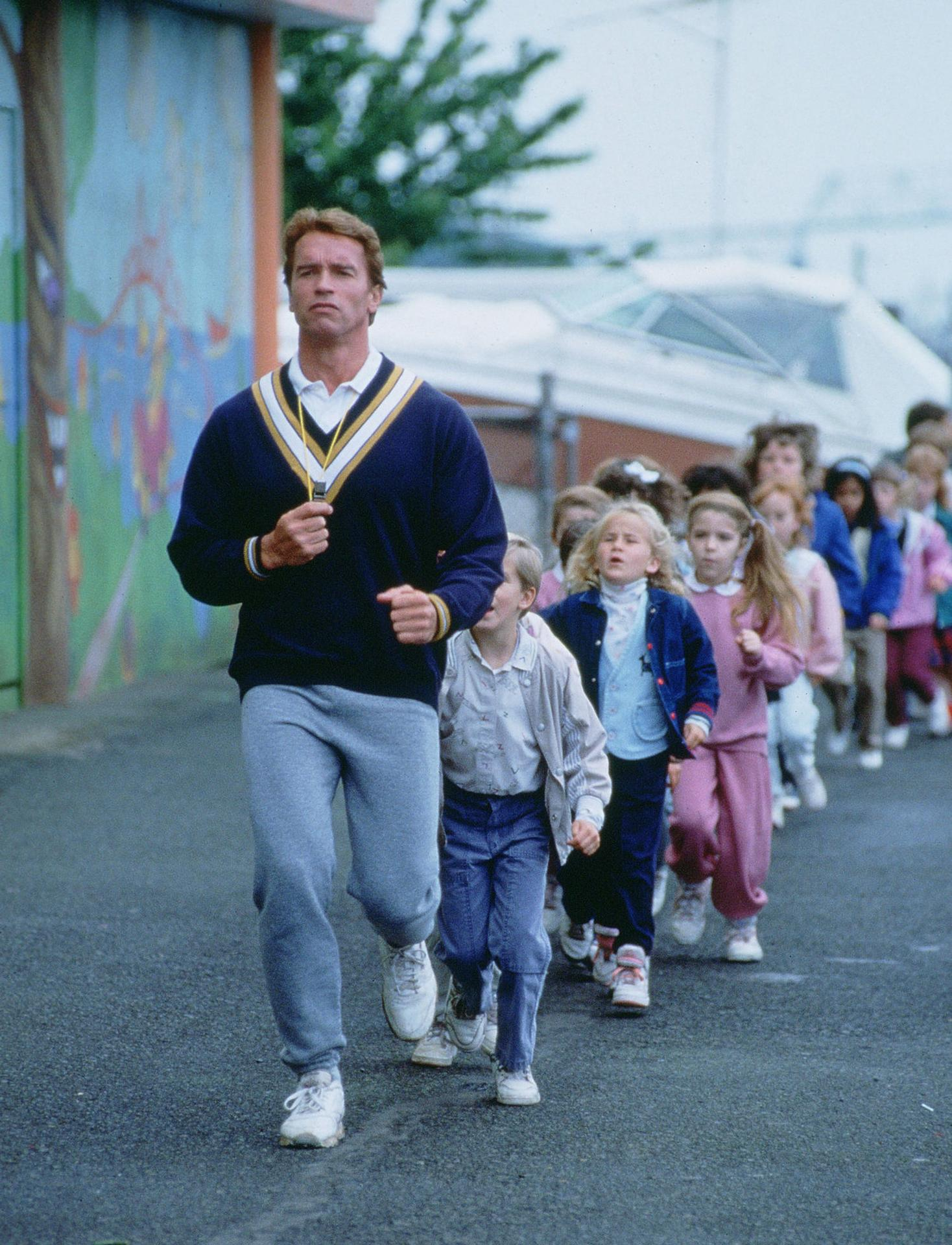 Arnold Schwarzenegger läuft mit einer Reihe von Kindern auf einem Schulhof.