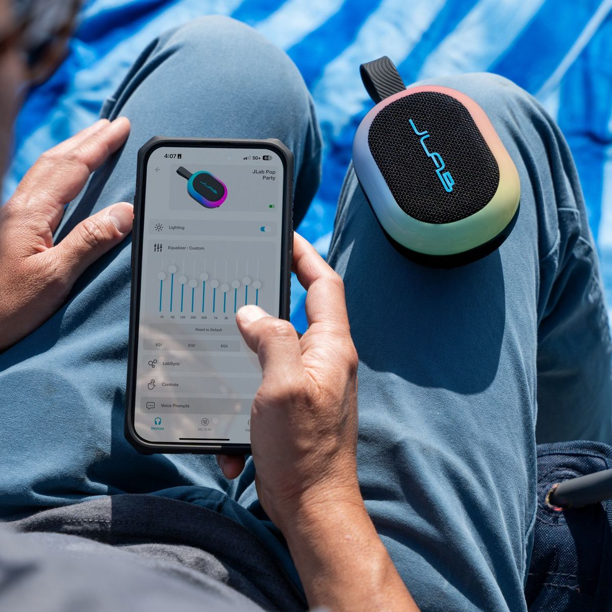 Person benutzt Handy mit JLab App zur Steuerung des bunten Lautsprechers im Schoß.