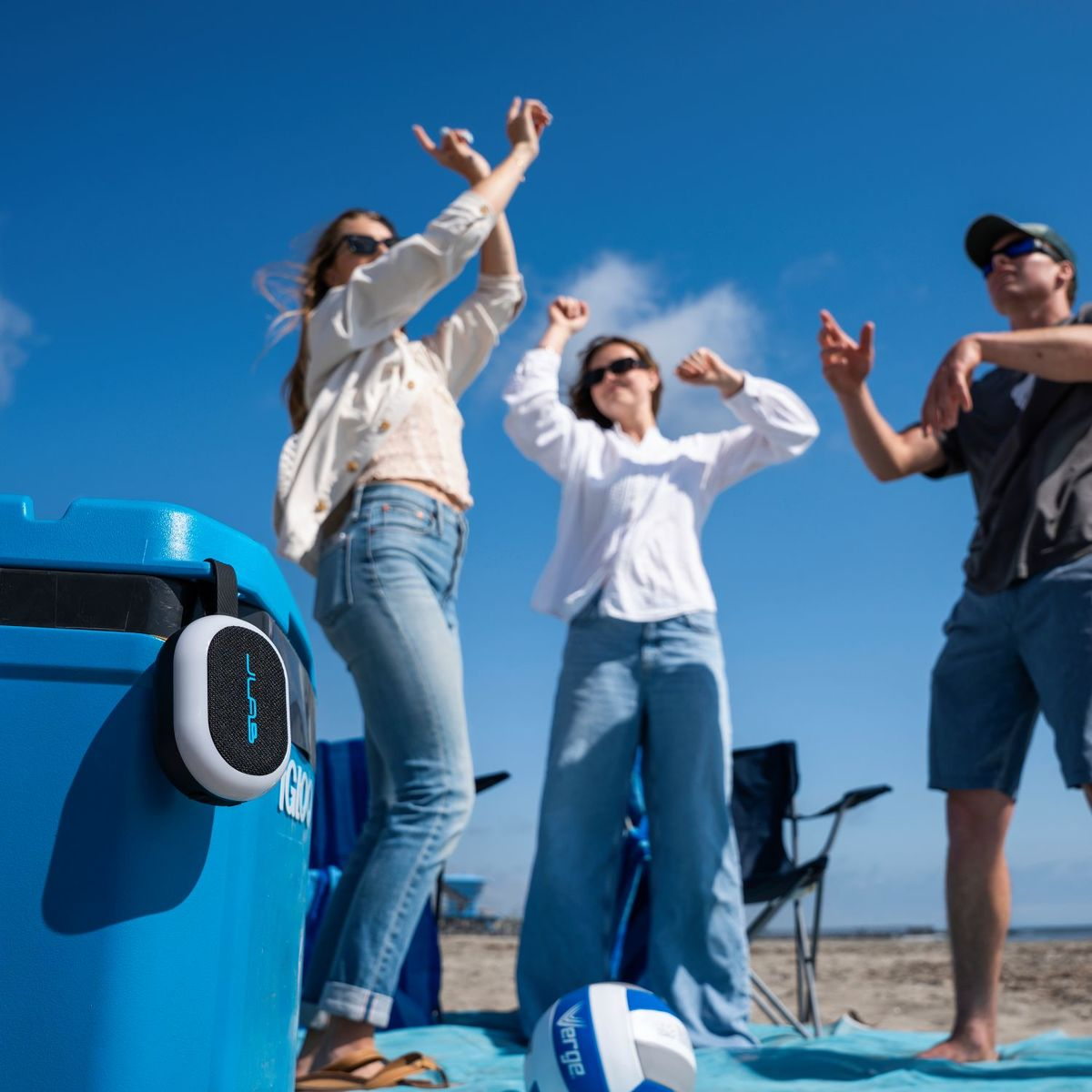 Drei Personen tanzen am Strand neben einer blauen Kühlbox und einem Volleyball.