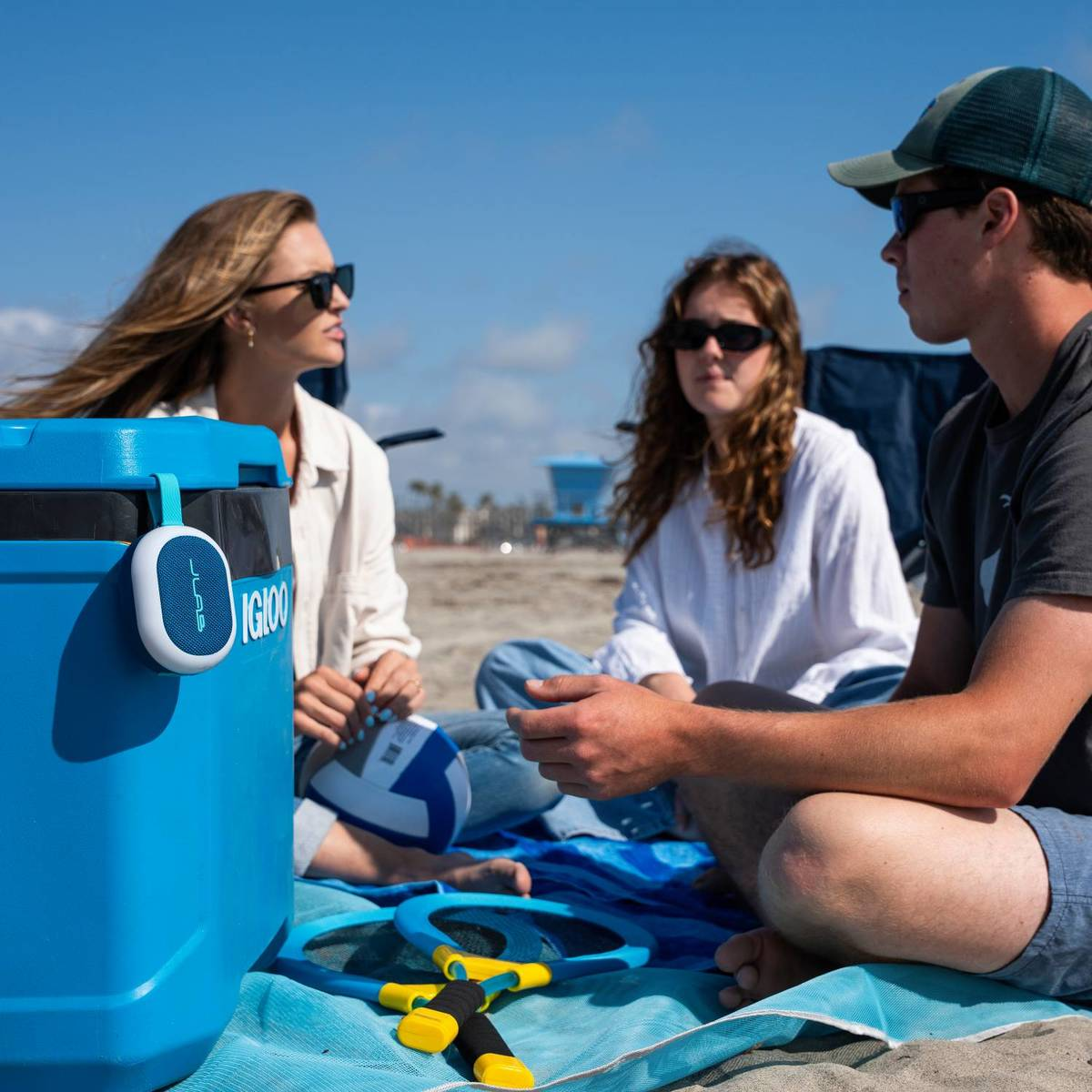 Drei Personen sitzen am Strand neben einer blauen Kühlbox. Ein kleiner Lautsprecher hängt an der Kühlbox.