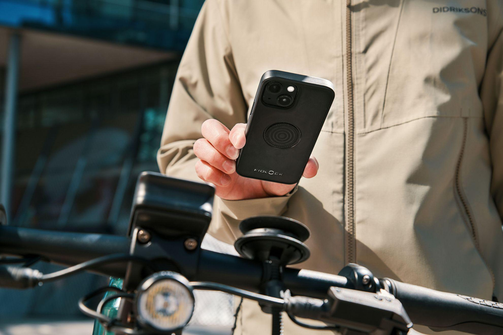 Person hält Handy über Fahrradlenker. Beige Jacke, Gebäude im Hintergrund.