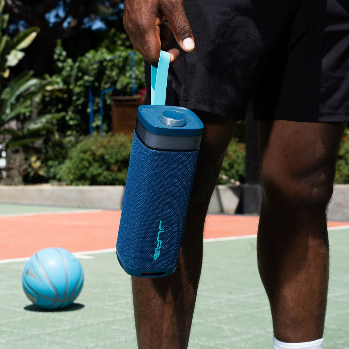 Ein Mann hält eine blaue Lautsprecherbox mit einem hellblauen Riemen. Ein blauer Basketball liegt daneben.