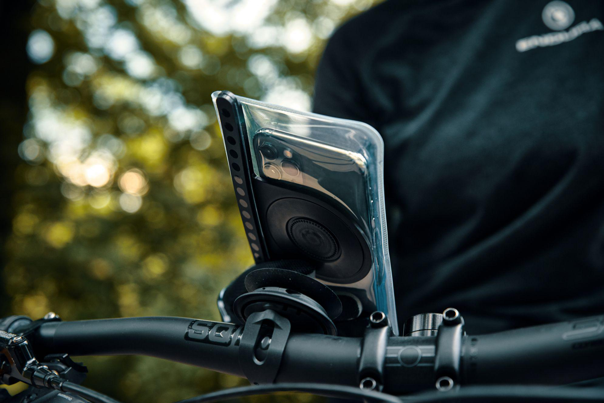 Telefon in klarer Hülle auf schwarzem Fahrradlenker montiert, draußen, verschwommener grüner Hintergrund.