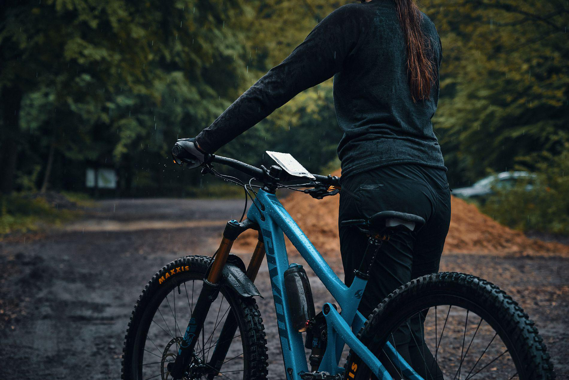 Eine Person steht im Regen neben einem blauen Mountainbike auf einem schlammigen Weg.