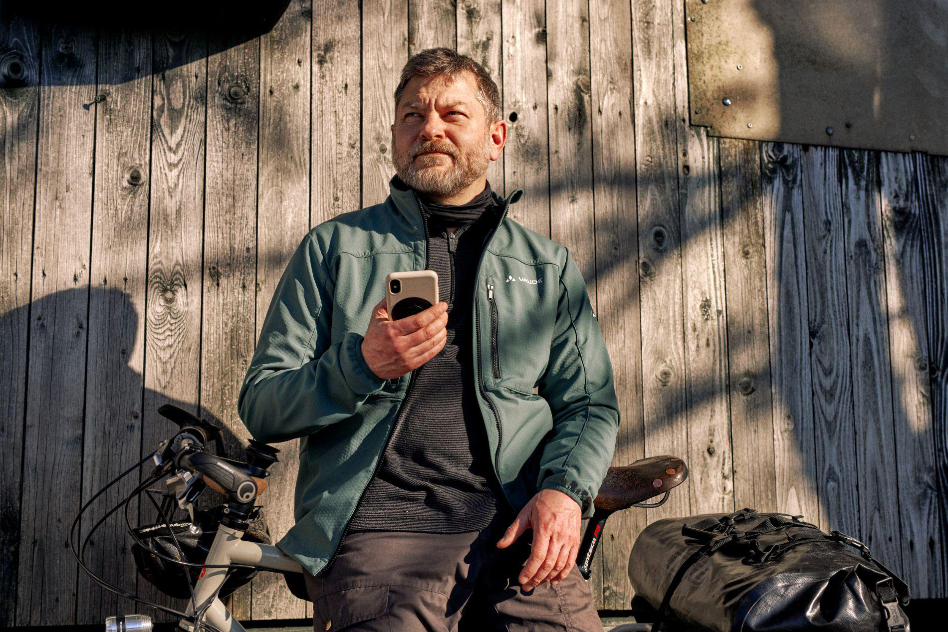 Ein Mann mit Bart steht neben einem Fahrrad vor einer Holzwand und hält ein Handy.