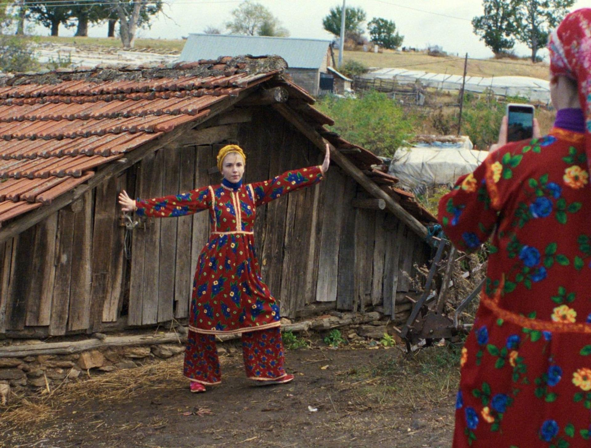 Frau in traditionellem Kleid tanzt vor Holzhütte, eine andere Person filmt mit Smartphone.