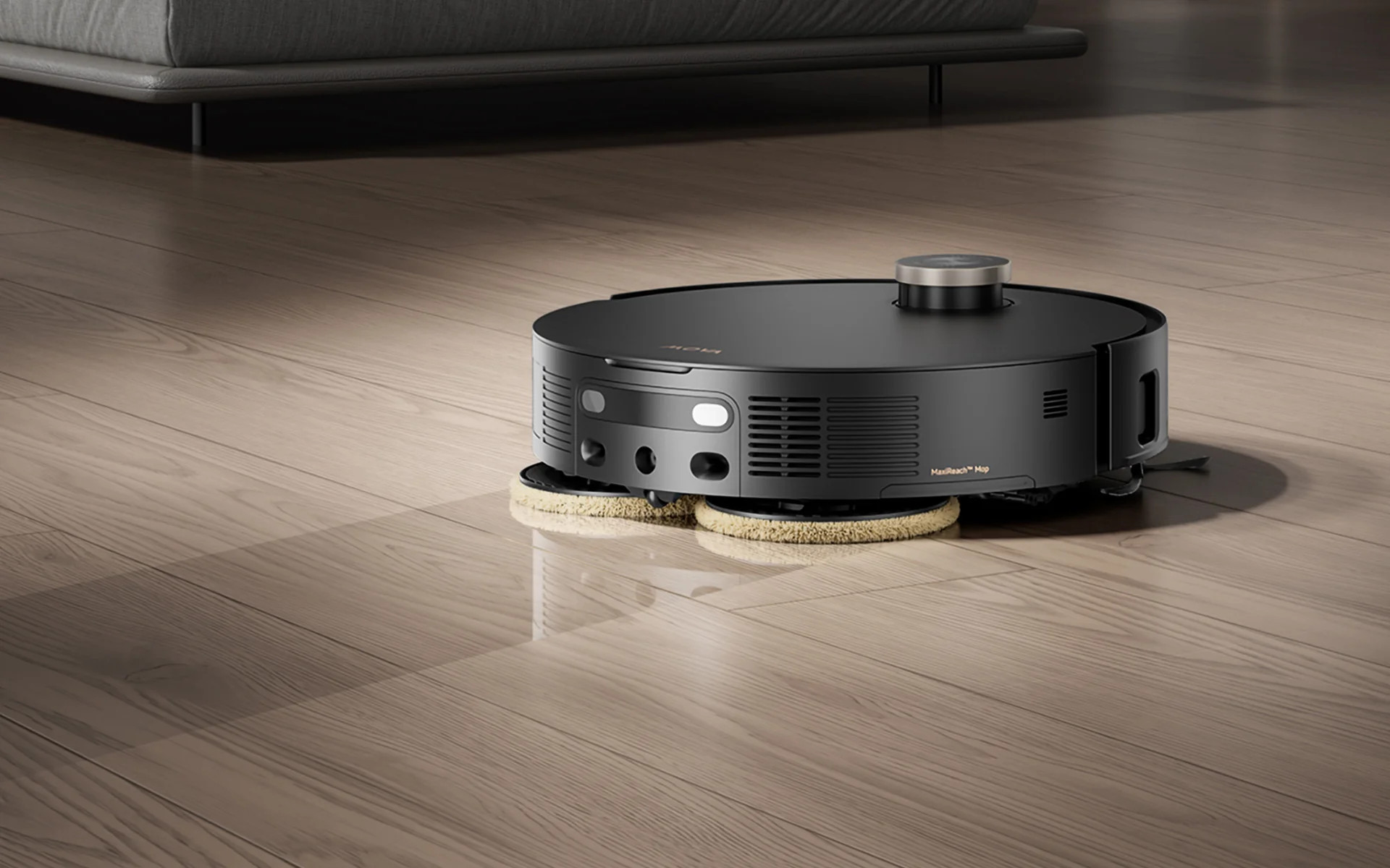 Czarny robot sprzątający z obrotowymi szczotkami czyści drewnianą podłogę.