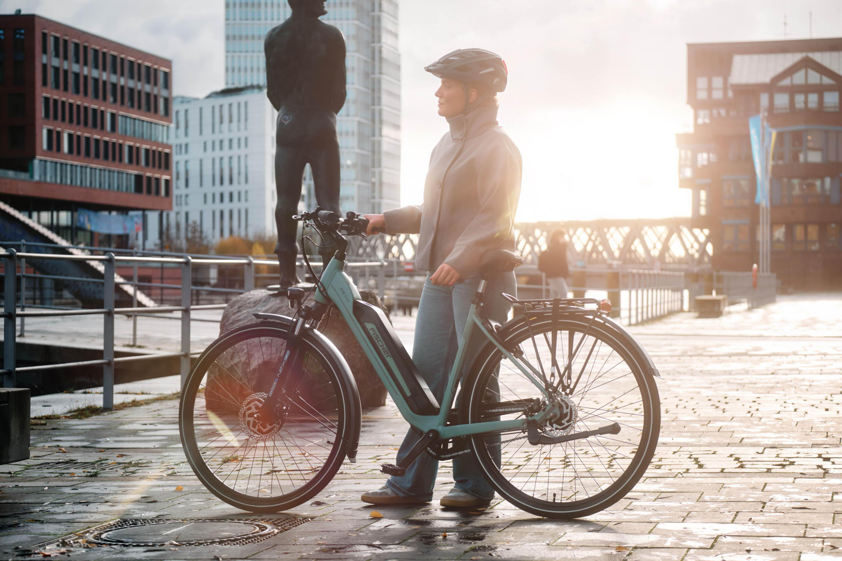 Eine Frau mit Helm steht neben einem Fahrrad auf einem nassen Gehweg mit Gebäuden im Hintergrund.