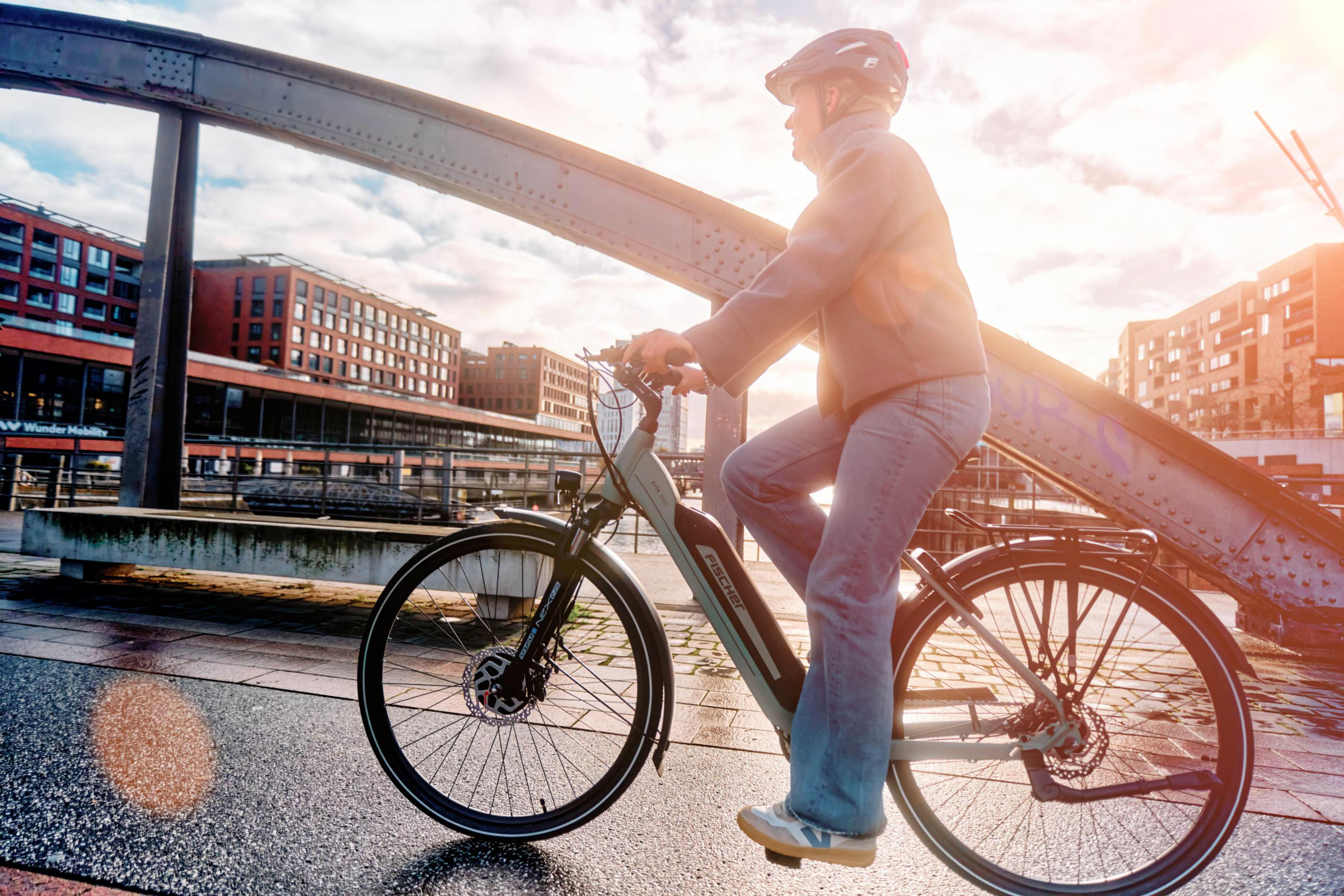 Person fährt Fahrrad auf Brücke mit Gebäuden im Hintergrund.